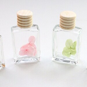 May include: Four small, clear glass bottles with wooden caps, each containing a different colored dried flower: white, pink, green, and blue. The bottles are square-shaped and arranged in a row, showcasing the delicate floral arrangements.