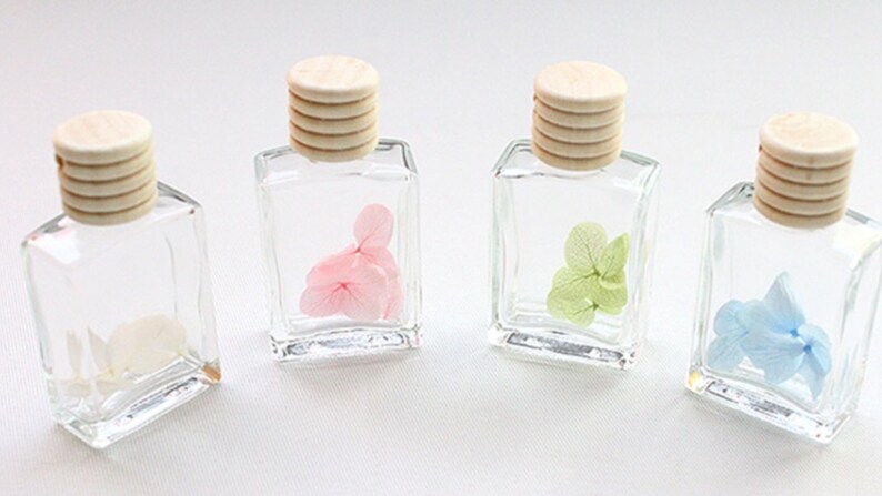 May include: Four small, clear glass bottles with wooden caps, each containing a different colored dried flower: white, pink, green, and blue. The bottles are square-shaped and arranged in a row, showcasing the delicate floral arrangements.