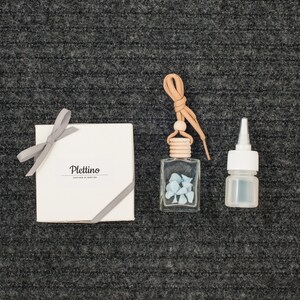 May include: An assortment of fragrance products, including a small clear glass bottle with blue stones, a small white bottle, and a white box tied with a gray ribbon. The box has the word "Plettino" printed on it.