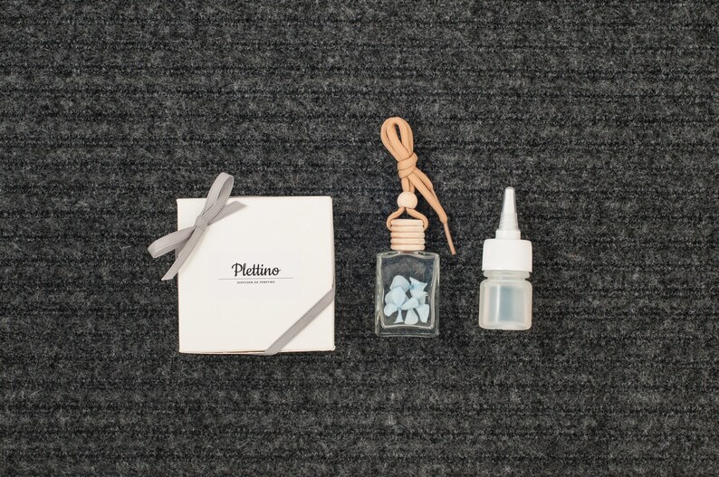May include: An assortment of fragrance products, including a small clear glass bottle with blue stones, a small white bottle, and a white box tied with a gray ribbon. The box has the word "Plettino" printed on it.