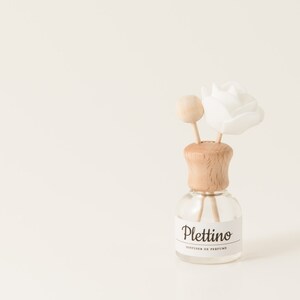 May include: A clear glass diffuser bottle with a wooden cap and a white flower and wooden stick inserts. The bottle has a label that reads "Plettino, Diffuser de Parfum". The diffuser is set against a cream-colored background.