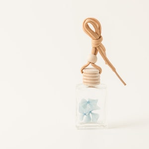 May include: A clear glass aromatherapy diffuser bottle with a light blue floral arrangement inside. The bottle has a wooden cap and a tan cord for hanging. The background is a solid off-white color.