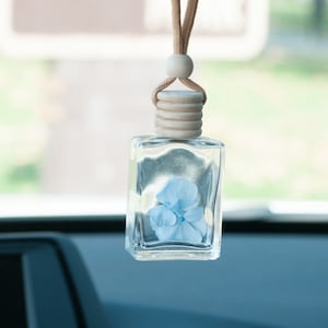 May include: A clear, square glass car air freshener with a light blue flower inside. The wooden top and string are attached to a wooden ball for hanging. The background is blurred.
