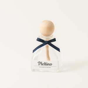 May include: A clear glass diffuser bottle with a wooden stopper and a navy blue ribbon. The bottle has a label that reads "Plettino, Diffuser of Perfume". The diffuser is designed to release fragrance into a room.