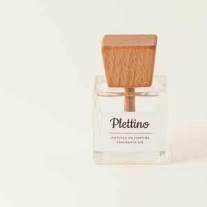 May include: A clear glass fragrance oil diffuser with a light brown wooden top. The label reads "Plettino, Diffuser de Perfume, Fragrance Oil". The diffuser is square-shaped and sits on a white background.