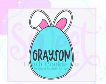 May include: A blue Easter egg cookie cutter with bunny ears. The egg has the name "GRAYSON" written on it.
