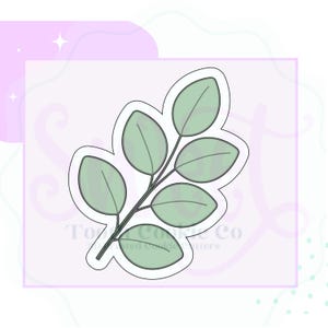 May include: A light green eucalyptus leaf cookie cutter with a white outline. The leaves are oval-shaped and arranged along a central stem. The cookie cutter is on a light pink background with a white border.