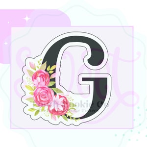 May include: A black cookie cutter in the shape of the letter G with a floral design on the left side. The flowers are pink and white with green leaves.