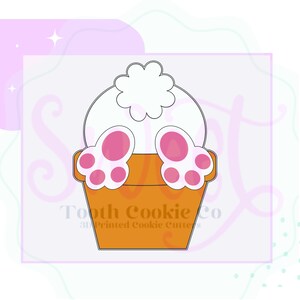 May include: A cookie cutter in the shape of a bunny's bottom and paws peeking out of a flower pot. The pot is orange, the bunny's bottom is white, and the paws are pink. The text "Tooth Cookie Co" is at the bottom.