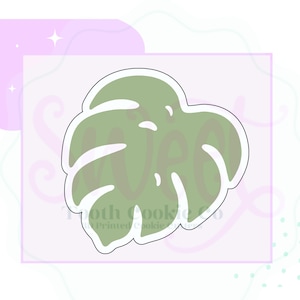 May include: A green monstera leaf cookie cutter with a white outline. The cutter is shaped like a large leaf with a pointed tip and a rounded base.