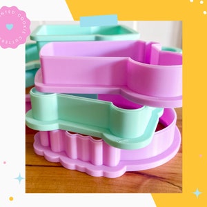 May include: Three 3D printed cookie cutters stacked on top of each other. The top cutter is a rectangle, the middle cutter is a light green paw print, and the bottom cutter is a light purple scalloped shape. The cutters are made of plastic and are designed for baking.