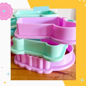 May include: Three 3D printed cookie cutters stacked on top of each other. The top cutter is light green, the middle cutter is light purple, and the bottom cutter is a darker shade of purple. The cutters are shaped like a heart, a star, and a flower. The text "Sweet Tooth cookie co." is visible at the bottom of the image.