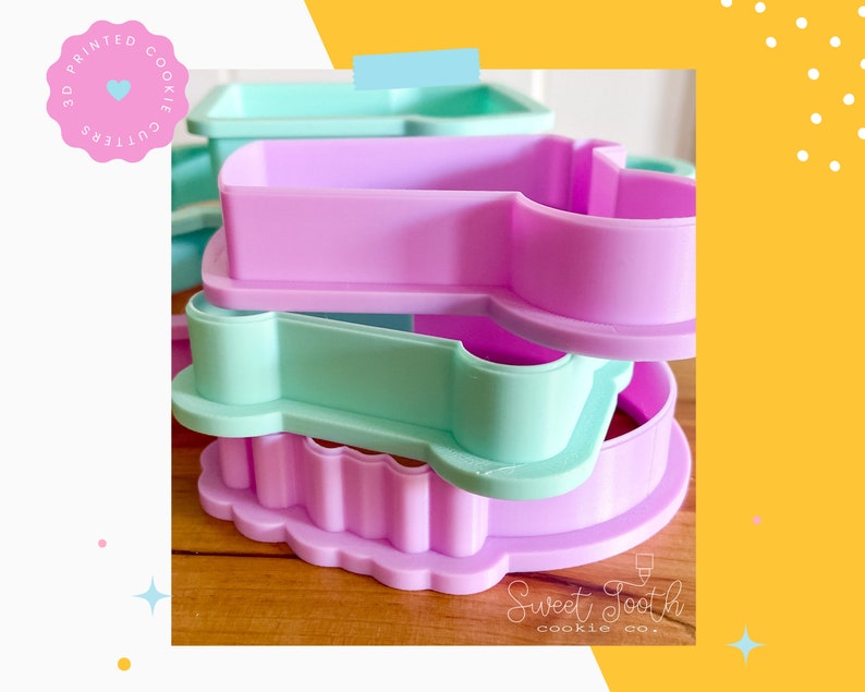 May include: Three 3D printed cookie cutters stacked on top of each other. The top cutter is light green, the middle cutter is light purple, and the bottom cutter is a darker shade of purple. The cutters are shaped like a heart, a star, and a flower. The text "Sweet Tooth cookie co." is visible at the bottom of the image.