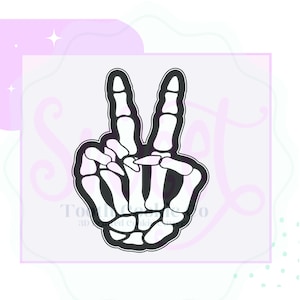 May include: A black and white skeleton hand cookie cutter in a peace sign gesture. The design is outlined in black, with the bone structure detailed in white. The background is a light lavender color with decorative shapes.