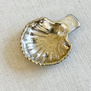 Op de afbeelding: Een kleine, zilverkleurige schelpvormige schaal met een getextureerd oppervlak. De schaal heeft een geschulpte rand en een klein handvat. De schaal is waarschijnlijk van metaal en ligt op een lichtgekleurde stof.