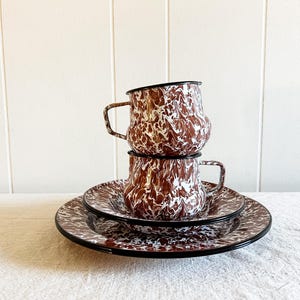 May include: A set of brown and white speckled enamelware dishes. The set includes two cups and a saucer. The cups and saucer have a black rim.