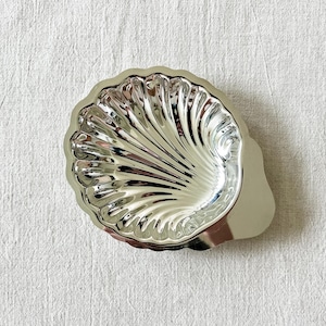 Könnte beinhalten: Eine polierte, silberfarbene Metallschale in Form einer Jakobsmuschel. Die Schale hat eine spiegelnde Oberfläche mit tiefen, strahlenförmigen Rillen, die die natürliche Textur einer Muschel nachahmen, und verfügt über einen kleinen, glatten Griff.