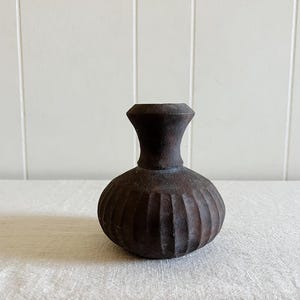 May include: A small, dark brown vase with a rounded, bulbous body and a narrow neck. The exterior surface features a series of vertical, carved ridges that extend from the base to the shoulder, creating a textured, matte appearance throughout.