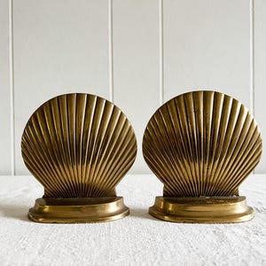 May include: Two brass seashell-shaped bookends. Each bookend features a fan-like design with vertical lines radiating from the base. The bookends are a warm gold color and sit on a white surface.