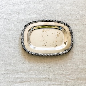 May include: A small, oval-shaped silver-toned metal tray featuring a decorative twisted rope-style border. The center of the tray has a smooth, reflective surface with visible dark spots and signs of natural oxidation throughout the metal.