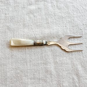 May include: A vintage serving fork with a mother-of-pearl handle and a silver-toned, three-pronged head. The handle is rectangular and the metal has decorative detailing. The fork is set against a neutral linen background.