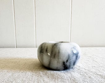 May include: A white marble bowl with gray veining. The bowl is round and has a smooth, polished surface.