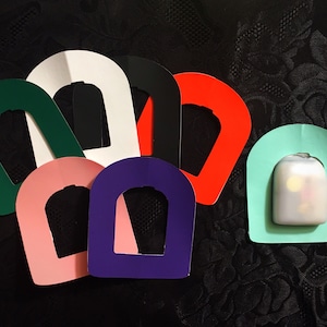 Può includere: Un set di sette forme di carta colorate, tagliate a forma di arco. Le forme sono di vari colori, tra cui verde, bianco, nero, rosso, rosa, viola e azzurro. Le forme sono disposte a semicerchio, con la forma azzurra in basso.