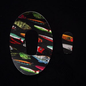 Op de afbeelding: Een ovale sticker in zwart en wit met een kleurrijk patroon van vislokken. De sticker heeft een uitsparing in het midden. Een kleinere, vergelijkbare sticker wordt rechts getoond.