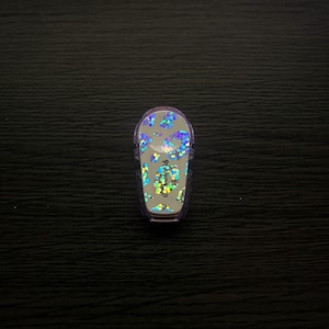 May include: A clear, plastic insulin pump with a glittery, iridescent leopard print design. The pump is small and oval-shaped.