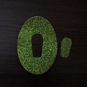 May include: Two glittery green shapes on a dark wood surface. One is a large oval with a cutout, and the other is a smaller, rectangular shape. The glitter adds a sparkling texture to the objects.