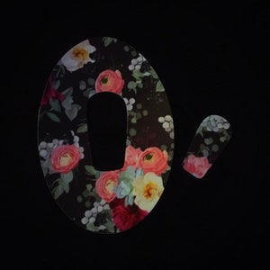 May include: A floral patterned sticker for a medical device. The sticker is shaped like a large oval with a smaller oval cut out of the center. The sticker has a black background with pink, red, and yellow flowers.