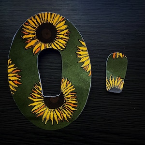 Puede incluir: Una pegatina ovalada verde con un diseño de girasol. La pegatina tiene un recorte en el centro para un sensor u otro dispositivo. La pegatina está rodeada de un fondo oscuro.