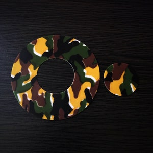 May include: Two round, flat, camouflage-patterned stickers. The larger sticker has a hole in the center. The smaller sticker is to the right of the larger sticker. Both stickers have a green, brown, and yellow camouflage pattern.