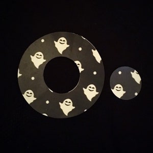 May include: Two black and white ghost-themed vinyl stickers. The larger sticker is a donut shape with a hole in the centre. The smaller sticker is a circle. Both stickers feature white ghosts with smiling faces on a black background.
