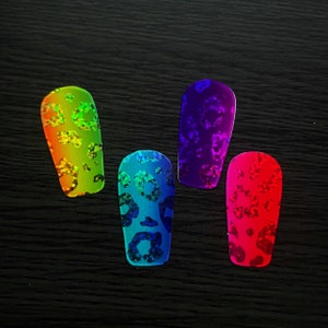 Puede incluir: Cuatro pegatinas de uñas iridiscentes con un diseño de estampado de leopardo. Las pegatinas son en tonos verde, azul, morado y rojo.