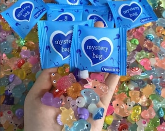 May include: A hand holds a collection of colorful, translucent miniature animal figurines. Surrounding the hand are several blue "mystery bag" packages with a heart-shaped design and the words "mystery bag". The figurines include ducks, fish, and other creatures.