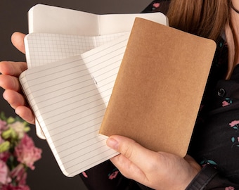 May include: A set of three small notebooks with rounded corners. One notebook has a plain brown cover, while the interior pages of the others feature white paper with either a grid pattern or horizontal lines for writing and note-taking purposes.