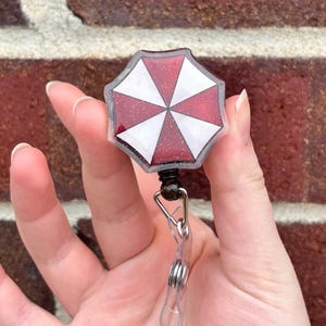 May include: A badge reel with a red and white umbrella design. The badge reel has a clear plastic strap and a black retractable cord. The umbrella design is an octagon shape with a glittery border.