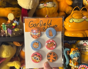 May include: A package of six Garfield buttons with various cartoon images of the orange cat. The buttons are round and feature different expressions and poses of Garfield. The package is held in a hand.