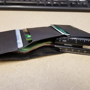 May include: A black leather wallet with multiple cards and cash inside. The wallet is open and a ruler is placed next to it. The ruler is marked in inches and centimeters.