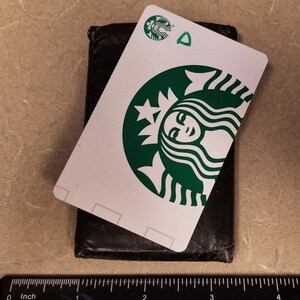 May include: A white Starbucks gift card with a green Starbucks logo featuring a woman's face. The card is sitting on top of a black leather wallet.