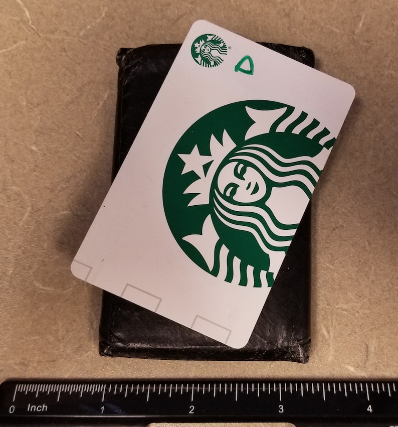 May include: A white Starbucks gift card with a green Starbucks logo featuring a woman's face. The card is sitting on top of a black leather wallet.