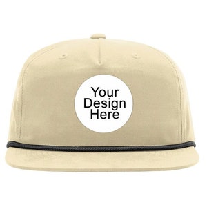Puede incluir: Una gorra de béisbol beige con ribete de cuerda negro y un círculo blanco con el texto "Your Design Here" en negro.