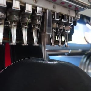 May include: Close-up of a black hat being embroidered with red and white stripes by a multi-needle embroidery machine.