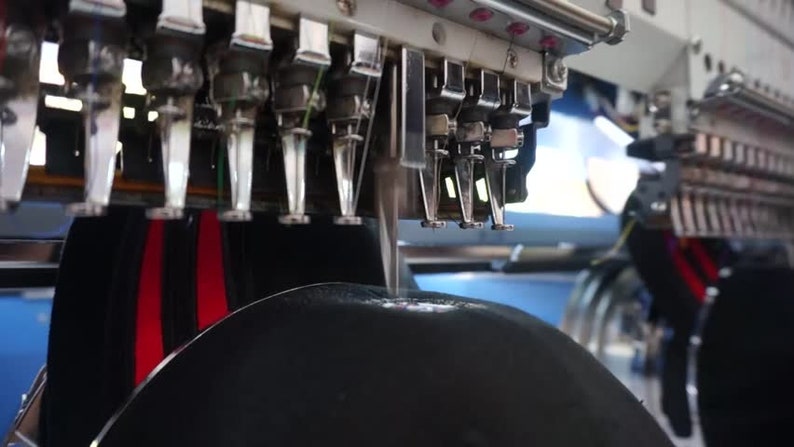 May include: Close-up of a black hat being embroidered with red and white stripes by a multi-needle embroidery machine.