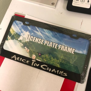 May include: Black license plate frame with a colorful image of a road and the text "LICENSE PLATE FRAME" and "ALICE IN CHAINS" printed on it.