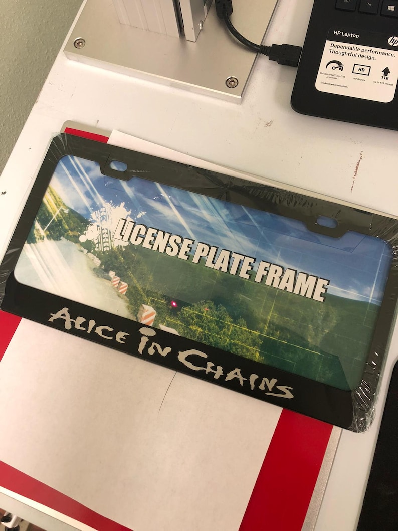 May include: Black license plate frame with a colorful image of a road and the text "LICENSE PLATE FRAME" and "ALICE IN CHAINS" printed on it.