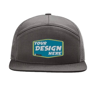 Op de afbeelding: Een grijze en zwarte truckercap met een mesh achterkant. De voorkant van de cap heeft een tealblauwe patch met de tekst "YOUR DESIGN HERE" in wit.