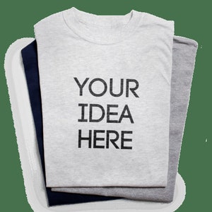 May include: Three folded t-shirts in gray, navy blue, and light gray. The top shirt is light gray with the text "YOUR IDEA HERE" printed in black. The shirts are neatly stacked, showcasing a simple, customizable design.