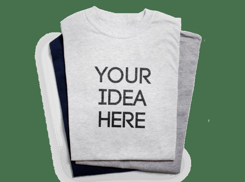 May include: Three folded t-shirts in gray, navy blue, and light gray. The top shirt is light gray with the text "YOUR IDEA HERE" printed in black. The shirts are neatly stacked, showcasing a simple, customizable design.